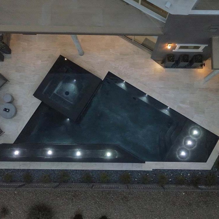 Drone view of a custom geometric infinity pool with integrated spa by California Pools, illuminated at night