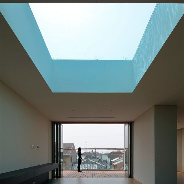 View of the living room of a modern design house, by Eastern Design Office, which has a window in the roof, through which you can see the swimming pool, and a wide opening onto a terrace on which a woman is standing