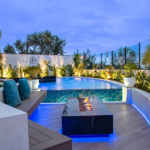 Impressive infinity pool project with contrasting raised spa, by Coastline Pools, in a minimalistly decorated garden, including a large outdoor lounge, sunken into the ground, with a gas firepit, at night