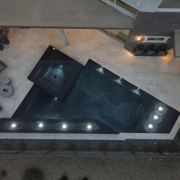 Drone view of a custom geometric infinity pool with integrated spa by California Pools, illuminated at night