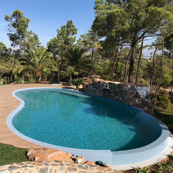 drone view of a kidney-shaped infinity pool with waterfall over decorative rocks, in a private garden surrounded by pine trees, with a decking terrace, by Piscinas Colombinas