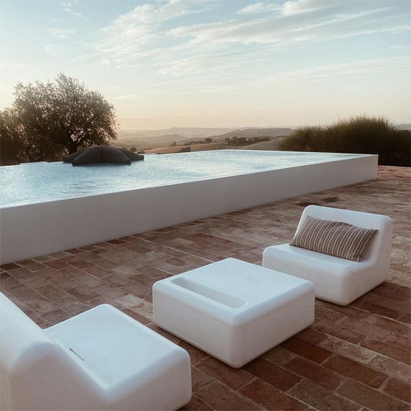 Infinity white pool, with a minimalist design, in a rustic space, in the middle of the countryside, from a project by Wespi, at sunset de Meuron Romeo Architetti, with some outdoor sofas and a modern design table, also in white, at sunset