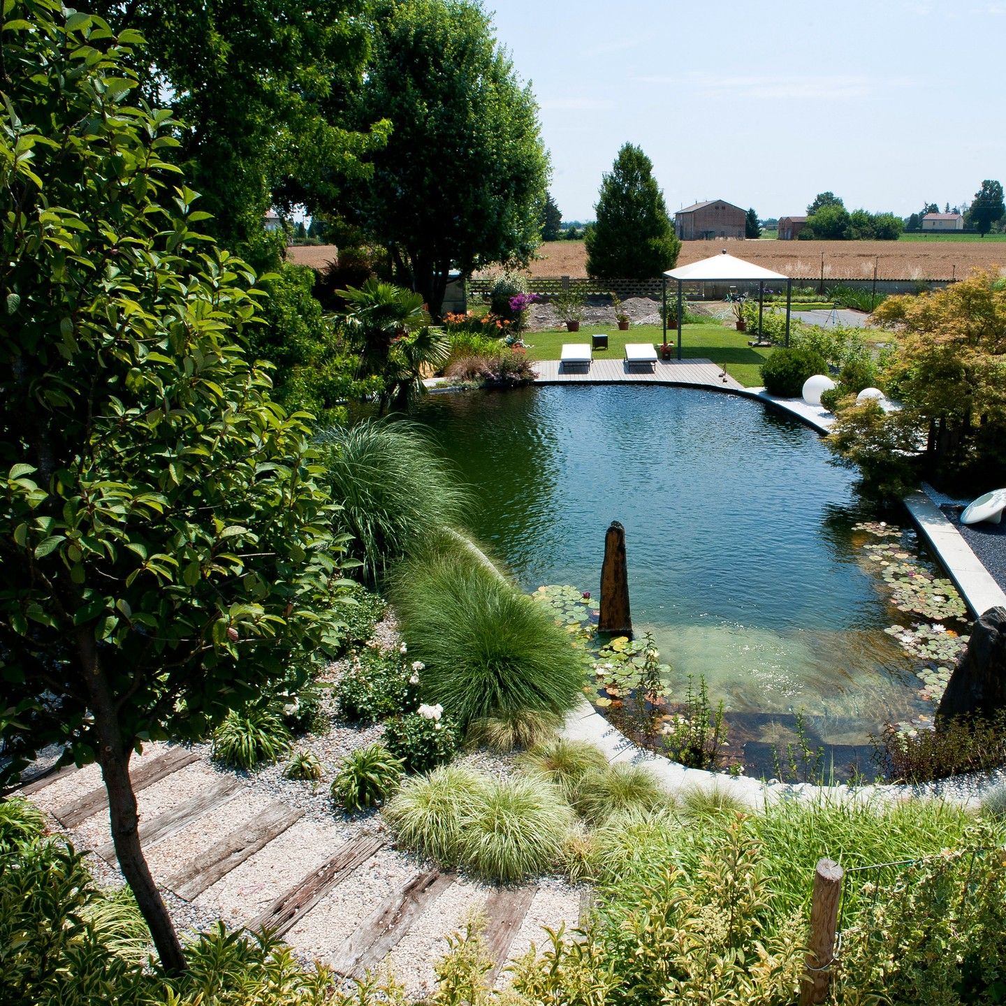 Custom natural pool in a huge garden, by Paesaggio Italiano, with a lot of plants and trees and a wooden decking as solarium