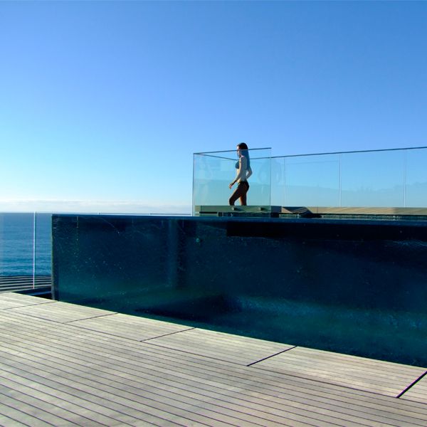 woman looking at the horizon from outside an above ground transparent infinity pool, by Aquatonic Architects, surrounded by a wooden deck