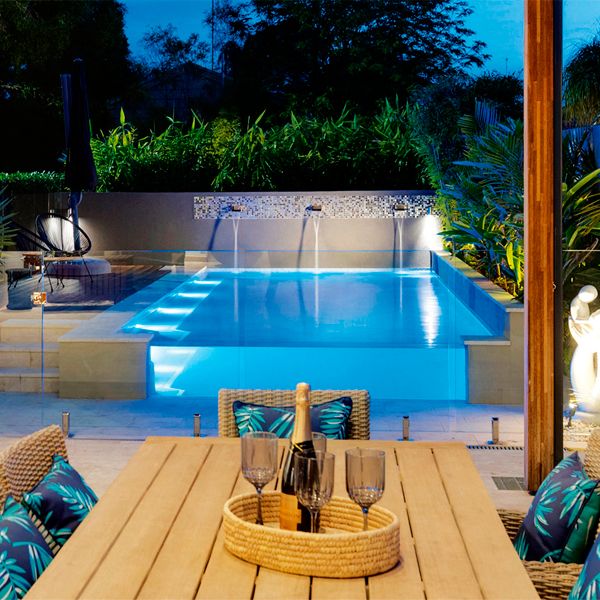 semi-inground private pool with a see-through wall by Jade Pools, lighted at night, viewed from the outdoor lounge area
