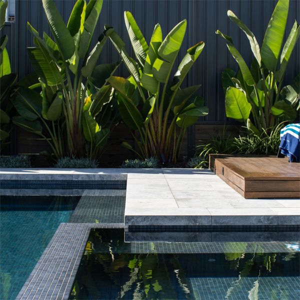 Detail of the interior coating of this elegant pool, in gray glass tile, the paved area, the garden area and wooden terrace, in a project by Splish Splash Pools