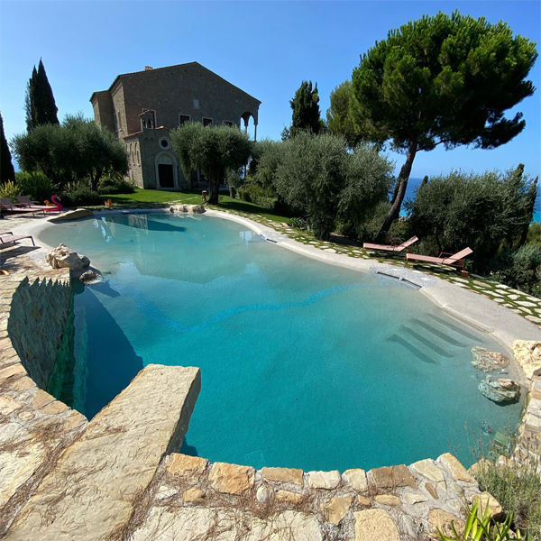 piscina estilo playa, realizada a medida por Piscine Sorgiva, rodeada de piedra, árboles y vegetación, en una casa de campo