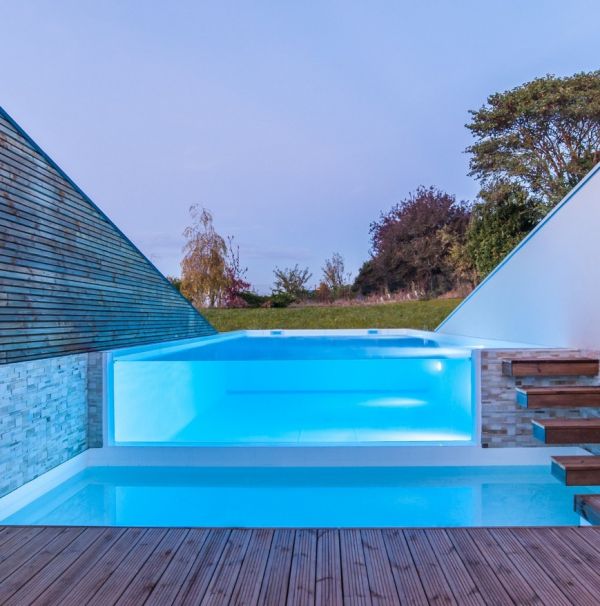 Preciosa y elegante piscina infinita, de diseño minimalista, con una pared transparente para ver su interior, por Piscinea, iluminada al atardecer