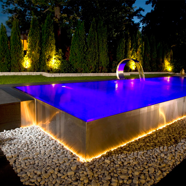 Piscina desbordante en acero inoxidable con cascada, también en acero, por Berndorf Pools, iluminada por la noche, en un jardín moderno y minimalista