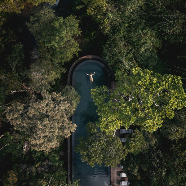 Vista dron de mujer nadando en piscina infinita en acero inoxidable, de diesño curvado, situada en medio de la jungla, dentro de un proyecto de hotel de Bill Bensley Architects