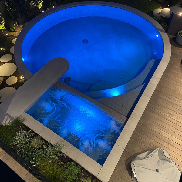 Piscina iluminada por la noche, de Exquisite Pools and Gardens, con un diseño único, en el que se integra un spa, y se combina el uso de líneas curvas y rectas, con formas circulares