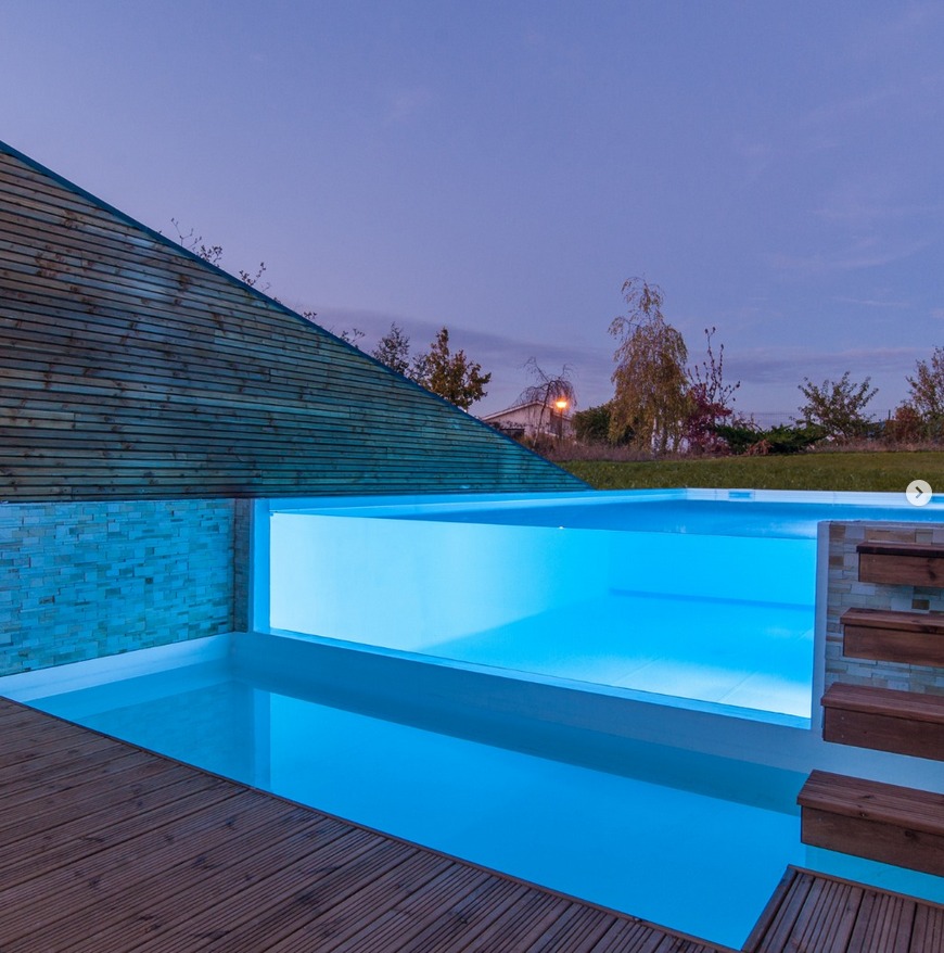 Preciosa y elegante mini piscina infinita, de diseño minimalista, con una pared transparente para ver su interior, por Piscinea, iluminada al anochecer