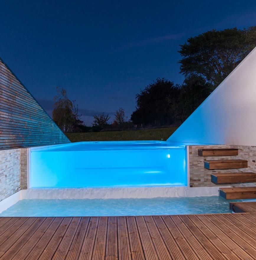 Preciosa y elegante piscina infinita, de diseño minimalista, con una pared transparente para ver su interior, por Piscinea, iluminada por la noche