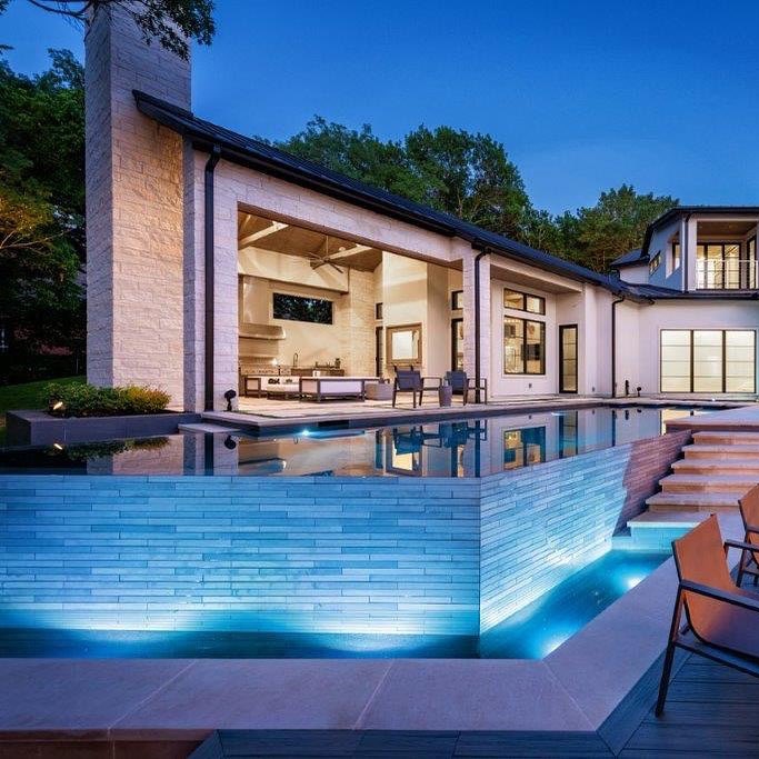 Piscina infinita de diseño en líneas rectas y ángulos, con revestimiento en color oscuro, por Pool Environments, iluminada por la noche