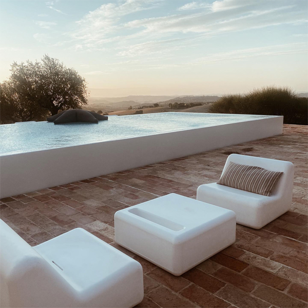piscina infinita, de diseño minimalista en color blanco, en un espacio rústico en medio del campo con unos sillones y una mesa de diseño moderno, también blancos, en un proyecto de Wespi de Meuron Romeo Architetti, al atardecer