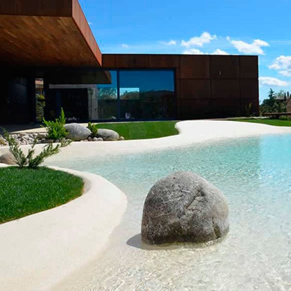 Piscine de sable, style plage, réalisée en Espagne, par Piscinas de Arena, et projet d'aménagement paysager complet combinant l'utilisation de la pierre, de la végétation et du bois dans l'environnement.