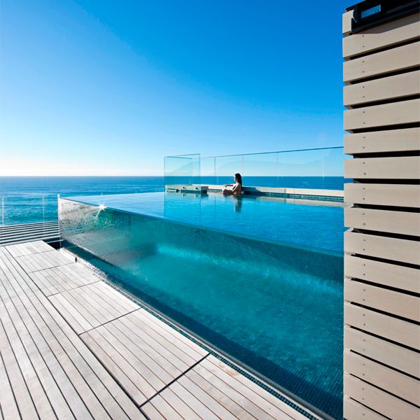 femme regardant l'horizon dans une piscine à débordement transparente, hors sol, par Aquatonic Architects, entourée d'une terrasse en bois