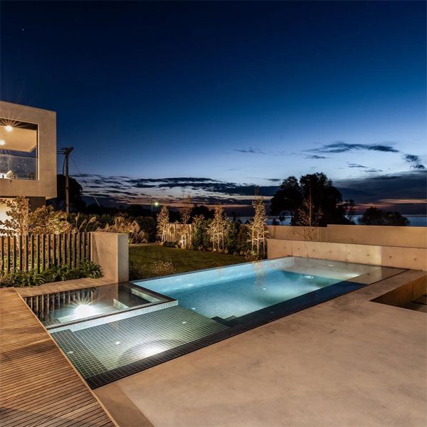 Élégante piscine à débordement avec spa intégré dans des tons sombres avec revêtement en mosaïque à l'intérieur combiné à de la pierre naturelle à l'extérieur, éclairée la nuit, par Coastal Pools and Spas